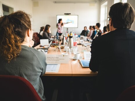 Personen an einem Konferenztisch, eine Frau hält eine Präsentation
