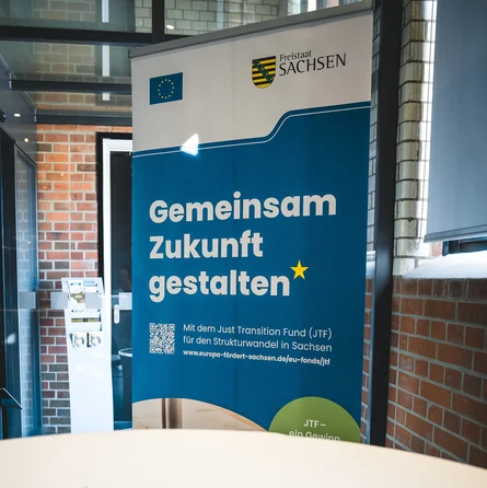 Ein blaues Roll-up mit der Aufschrift "Gemeinsam Zukunft gestalten" mit den Logos der EU und vom Freistaat Sachsen in einem Veranstaltungsraum