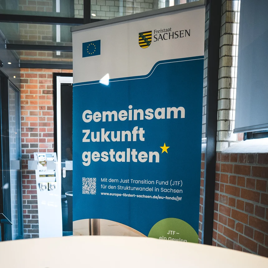 Ein blaues Roll-up mit der Aufschrift "Gemeinsam Zukunft gestalten" mit den Logos der EU und vom Freistaat Sachsen in einem Veranstaltungsraum