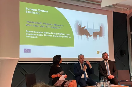 Podiumsdiskussion mit Staatsminister Schmidt und anderen Personen, im Hintergrund eine Präsentation zum JTF